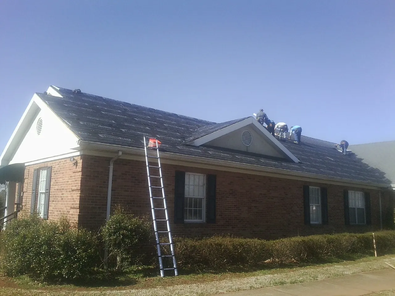 Crew tearing shingles off a brick ranch roof with a ladder set — Metal Roof Repair work by our crew in Brandamore