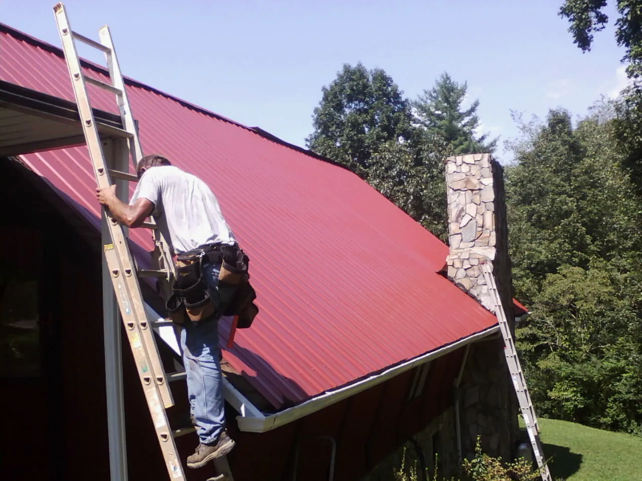 Roofer on a ladder beside a red metal roof and stone chimney — Soffit & Fascia Repair by Roofing Brothers in Brandamore